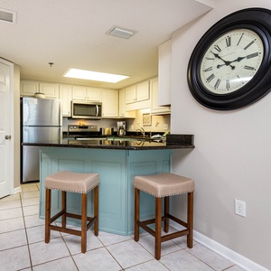 Bright kitchen area features a sleek island with bar stools, stainless appliances, and a stylish clock on the wall