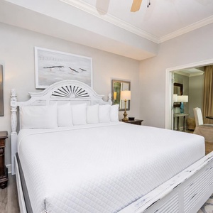The master bedroom features a stylish white king bed, a cozy lamp, and a lovely view of the beachfront through large windows