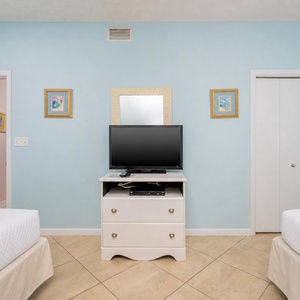 A light blue guest room features two double beds, a TV on a white dresser, and colorful art on the walls
