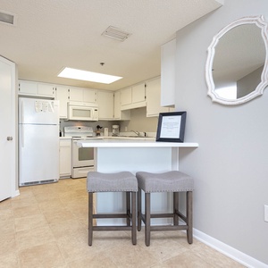 Bright, fully-equipped kitchen features white cabinetry, a breakfast bar with two stools, and a stylish mirror on the wall
