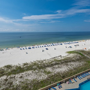 The view showcases a serene beach with white sand, colorful umbrellas, and a sparkling pool area below, inviting relaxation