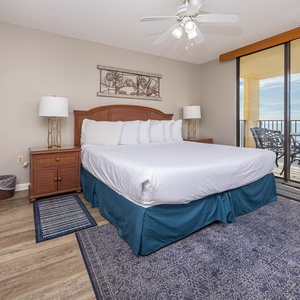 Bright master bedroom featuring a king bed, decorative lamps, and balcony access with seating for enjoying the beach view