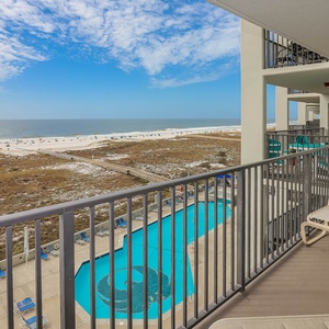Balcony view overlooking the pool area and the beach, featuring white sand and gentle waves under a bright blue sky