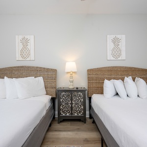 Two queen beds with wicker headboards and white linens are flanked by a decorative nightstand and pineapple-themed wall art