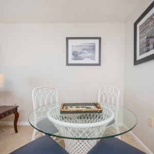 A round glass dining table with white wicker chairs is set against a neutral wall adorned with framed artwork