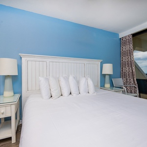 This guest bedroom features a plush king bed with white linens, a light blue accent wall, and a balcony view of the beach