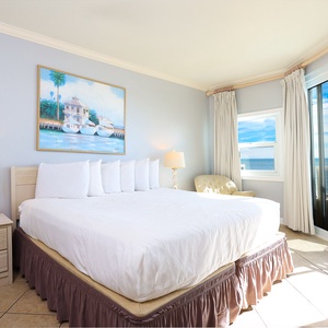 The master bedroom features a large bed with fluffy pillows, a cozy chair, and stunning beachfront views with balcony access