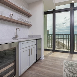 This wet bar features a sleek design with a wine cooler and glassware offering stunning beach views through large windows