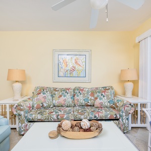 The living area features a colorful floral sofa, a light blue chair, and a view of the beach through sliding glass doors
