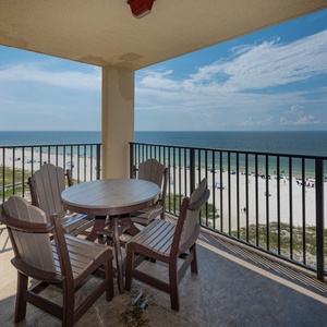 The balcony features a round table with wooden chairs, offering a serene view of the beach and calm waters beyond
