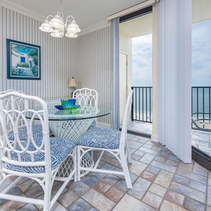Bright dining area features a glass table with blue-patterned chairs and a stunning view of the beach from the balcony