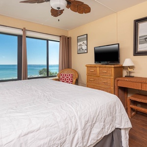 The master bedroom features a white quilted bed, a TV, and stunning beachfront views through large windows