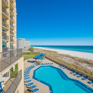 The balcony view showcases a sparkling pool area surrounded by lounge chairs, with a pristine beach and clear blue skies