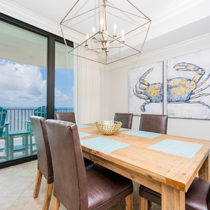 Dine at the wooden table surrounded by stylish chairs, with coastal artwork and a view of the inviting balcony
