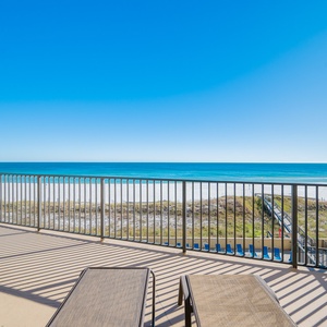 Relax on this large balcony with lounge chairs, enjoying the serene beach view and vibrant blue sky stretching over the water