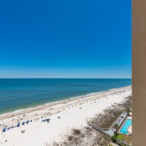 Balcony view showcasing a serene beach with white sand, colorful umbrellas, and gentle waves under a clear blue sky