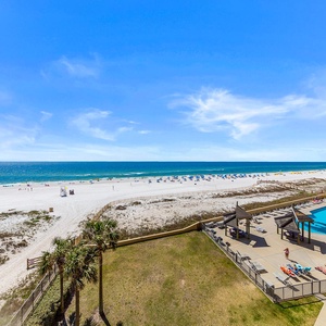 Expansive view of the beach with white sand, colorful umbrellas, and a pool area featuring lounge chairs and shaded pavilions