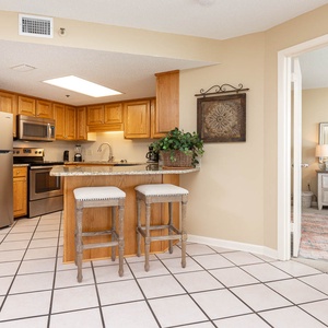 Kitchen area features wood cabinetry, granite countertops, and a breakfast bar with two stools, leading to a cozy bedroom