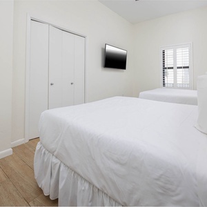 Bright guest bedroom featuring two double beds, a mounted TV, and a window with shutters, creating a welcoming atmosphere