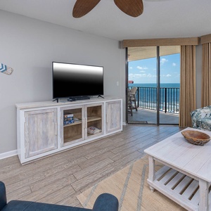 The living area features a comfortable sofa, a TV on a rustic cabinet, and large windows offering a breathtaking beach view
