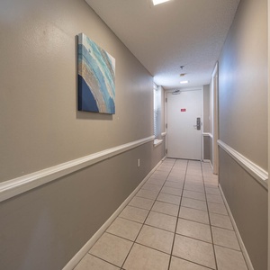 A serene hallway features light gray walls, tiled flooring, and a decorative blue artwork, leading to the entrance door