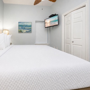 Bright and airy bedroom featuring a large king bed with plush white linens, a wall-mounted TV, and coastal artwork