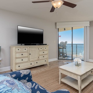 The living area features a cozy sofa, a TV on a light wood console, and expansive views of the beach