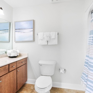 This bathroom features a stylish vanity with wooden cabinetry, tub-shower, fresh towels, and a serene coastal-themed artwork