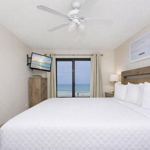 This bedroom features a cozy bed, a mounted TV, and a stunning beachfront view from the large window