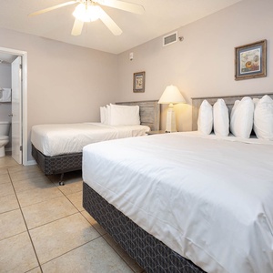 Bright guest room featuring two double beds with plush white bedding, a bedside lamp, and a door leading to a bathroom area