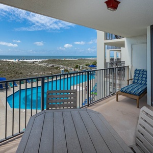 Balcony seating overlooks the pool area and offers a view of the beach with blue skies and distant beach umbrellas
