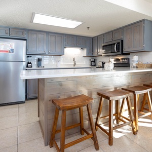 The kitchen features gray cabinetry, a white countertop, and wooden bar stools, creating a welcoming space