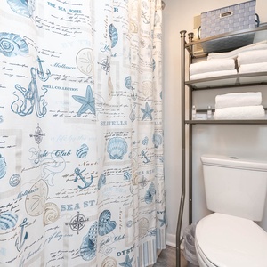 The bathroom features a beach-themed shower curtain, neatly arranged towels, and a clean, bright atmosphere for relaxation