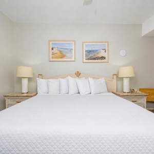 Rest in this spacious bedroom, featuring a large king bed, beach-themed artwork, and soft lighting for a relaxing atmosphere