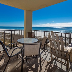 The balcony features a glass-top table with four chairs, offering a panoramic view of the tranquil beachfront and blue sky