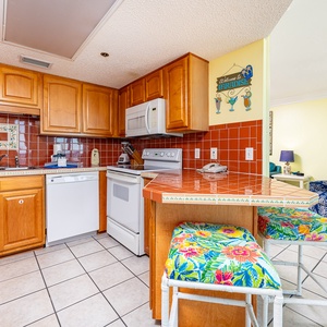 Kitchen with tiled breakfast bar, featuring wooden cabinetry and a cozy living area visible in the background