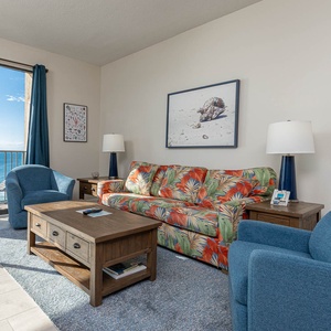 Living area features a floral sofa with queen sleeper, blue chairs, and expansive windows with a beautiful beach balcony view