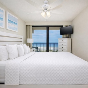 This bedroom features a plush white bed, a TV, and stunning beach front views through large sliding glass doors