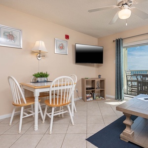 The living area features a cozy dining table, a wall-mounted TV, and stunning beach views through balcony doors