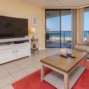 Living area features a large TV, cozy seating, and a view of the beach from the sliding glass doors leading to the balcony