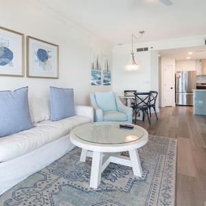 Relax in this sleek living space with cozy seating, a glass-top table, and coastal art to enhance your beach getaway