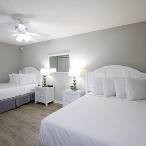 Bright guest bedroom featuring two double beds, white bedding, stylish nightstands, and a ceiling fan for comfort
