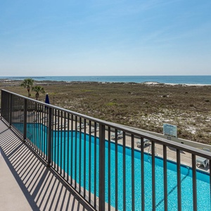 Balcony view overlooking the pool area and the tranquil beach, framed by lush greenery and a clear blue sky