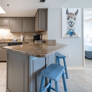 Modern kitchen features granite countertops and blue bar stools, with a whimsical llama painting adding a playful touch