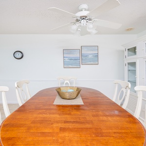 Bright dining area features a wooden table with light chairs, a decorative bowl, and coastal art enhancing the relaxed vibe
