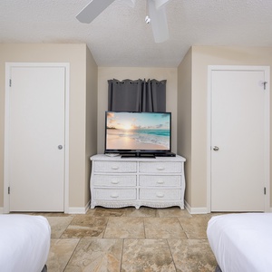 A cozy guest room features two double beds, a white dresser, and a TV displaying a serene beach scene