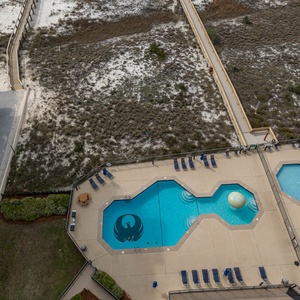 Aerial view showcases the pool area with vibrant blue waters, lounge chairs, and a shaded gazebo near the beach access path