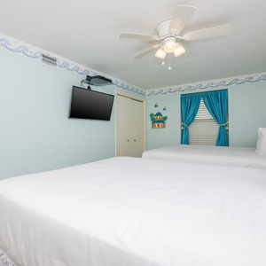 Bright guest room featuring two queen beds, a wall-mounted TV, and cheerful beach decor with vibrant blue curtains