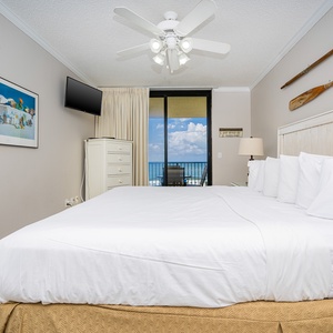 The master suite featuring a king bed, a mounted TV, and a balcony with a view of the beach and inviting blue skies