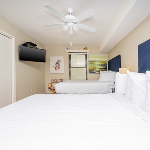 Guest bedroom features two queen beds with plush white bedding, a mounted TV, and colorful artwork against soft beige walls